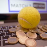 Auf einer Computertastatur liegt ein gelber Tennisball. Unter dem Ball liegen viele 1- und 2-Euro-Münzen.