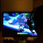 Ein Jugendlicher sitzt mit Kopfhörern vor dem PC. Im Hintergrund ist auf dem Bildschirm ein Videospiel zu sehen, das verschwommen erscheint.