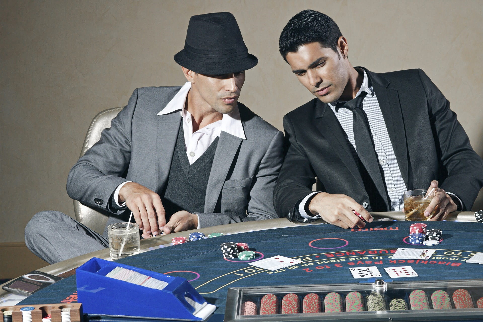 Zwei Männer im Anzug und Trinkgläsern sitzen an einem Pokertisch, auf dem Spielchips und einzelne Karten liegen.