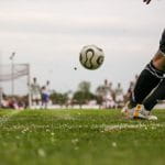 Zwei Beine und Fußball. Im Hintergrund weitere Fußballspieler, die verschwommen sind.