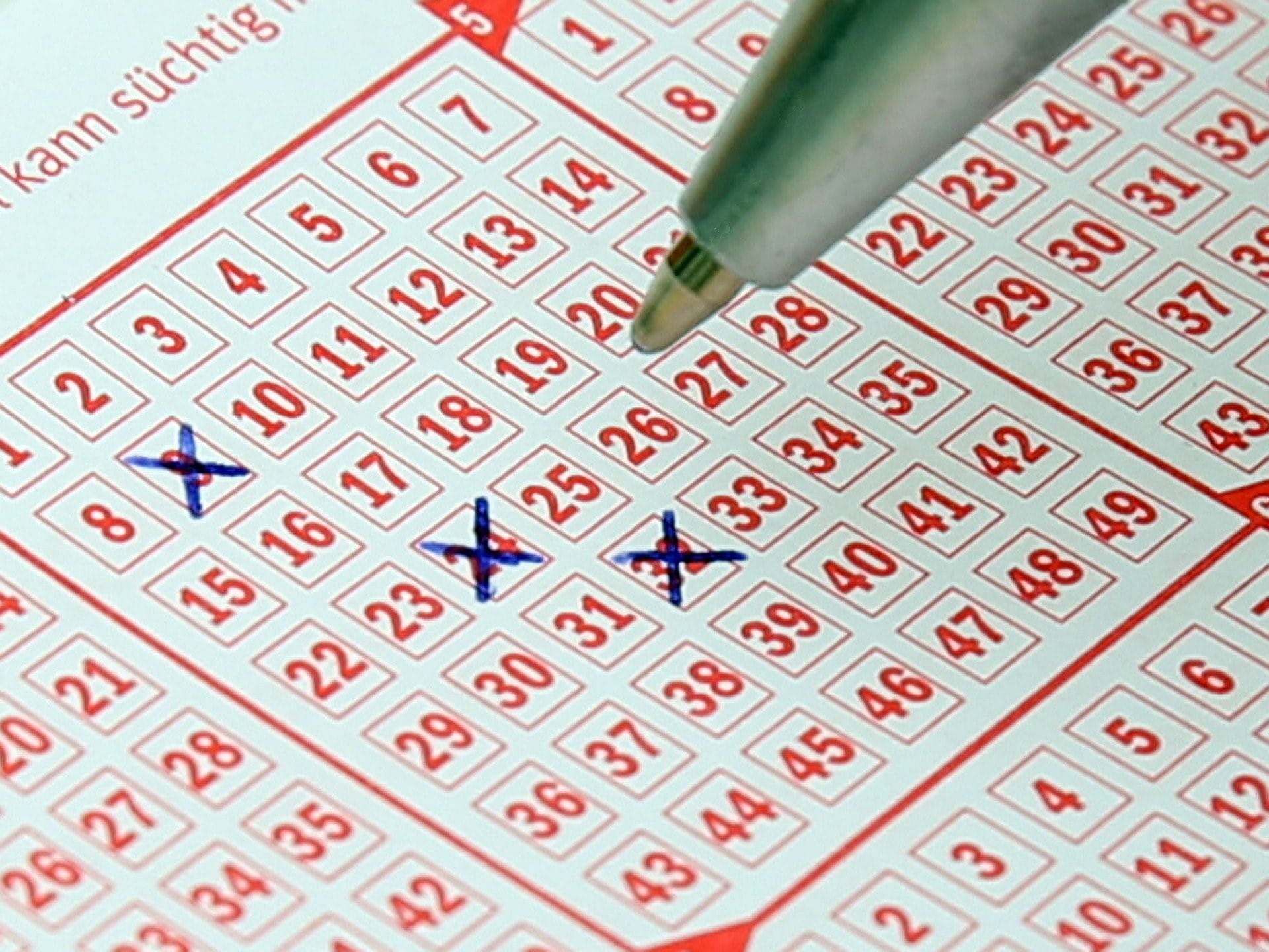 Ein roter Lottoschein, auf dem bereits drei Zahlen angekreuzt wurden. Ein Kugelschreiber schwebt über dem Lottoschein.