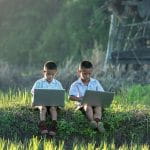 Zwei Kinder sitzen mit je einem Laptop auf dem Schoß in einer Wiese.