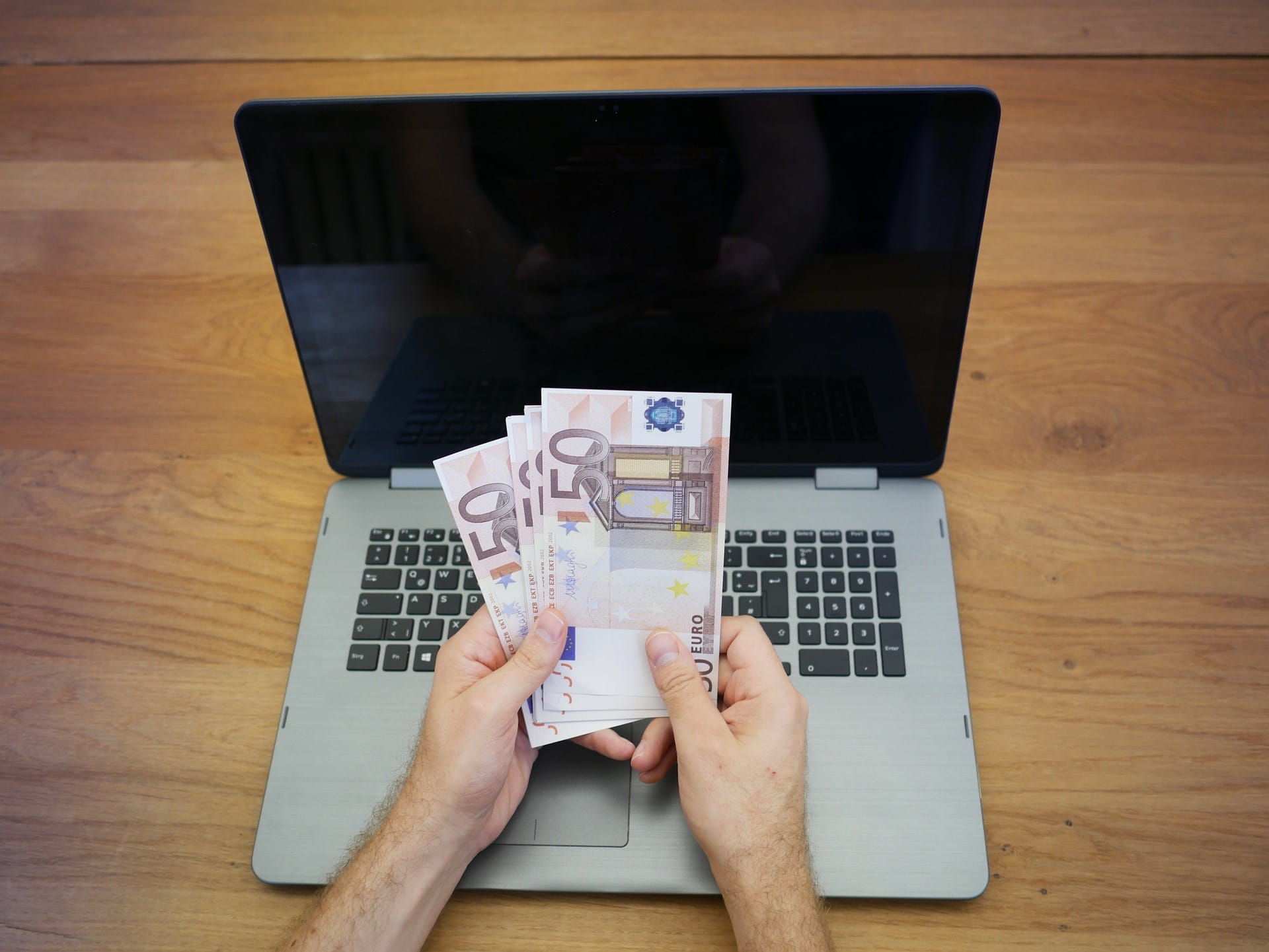 Ein Mann sitzt vor einem aufgeklappten Laptop und hält vier 50-Euro-Scheine in der Hand.