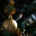 Eine goldene Weihnachtskugel mit Schleife und Tannenzweig verziert hängt am Baum.