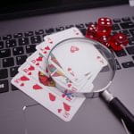 Auf einer Tastatur liegen Pokerkarten, vier rote Würfel und eine Lupe.
