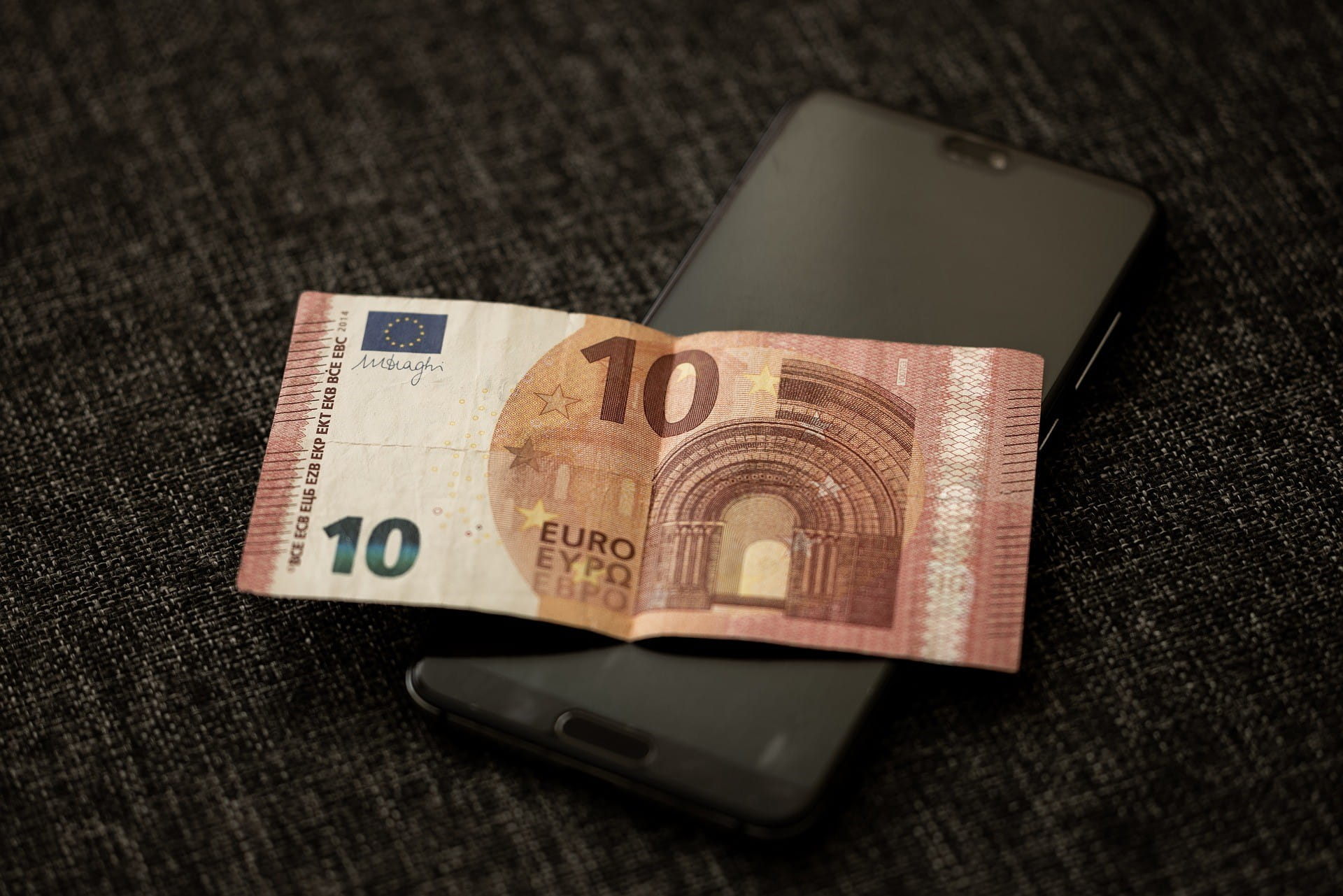 Auf einem abgedunkelten Smartphone liegt ein Zehn-Euro-Schein.