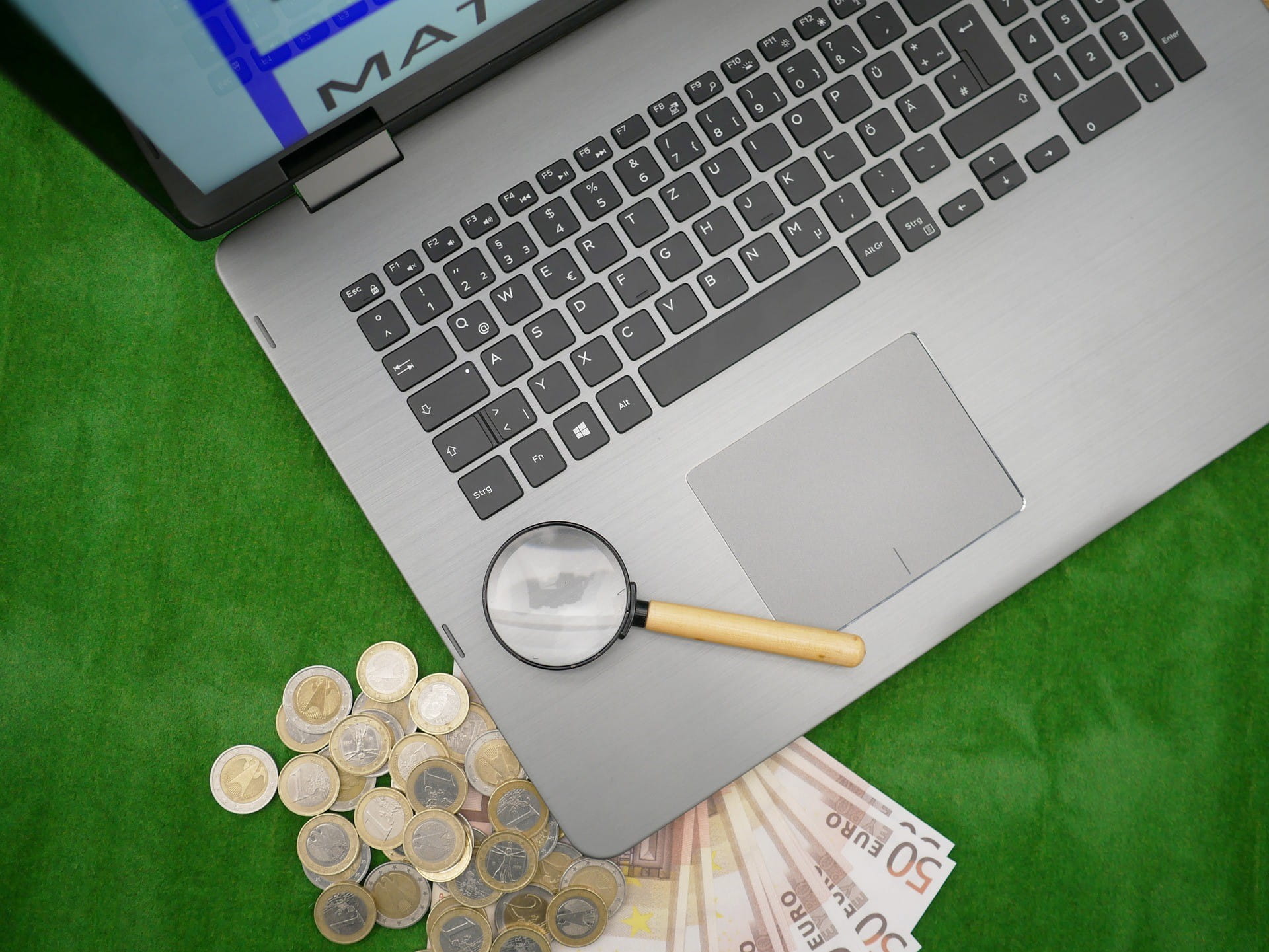 Auf einem Laptop liegt eine kleine Lupe, unter dem Laptop liegen Geldscheine und Geldmünzen.