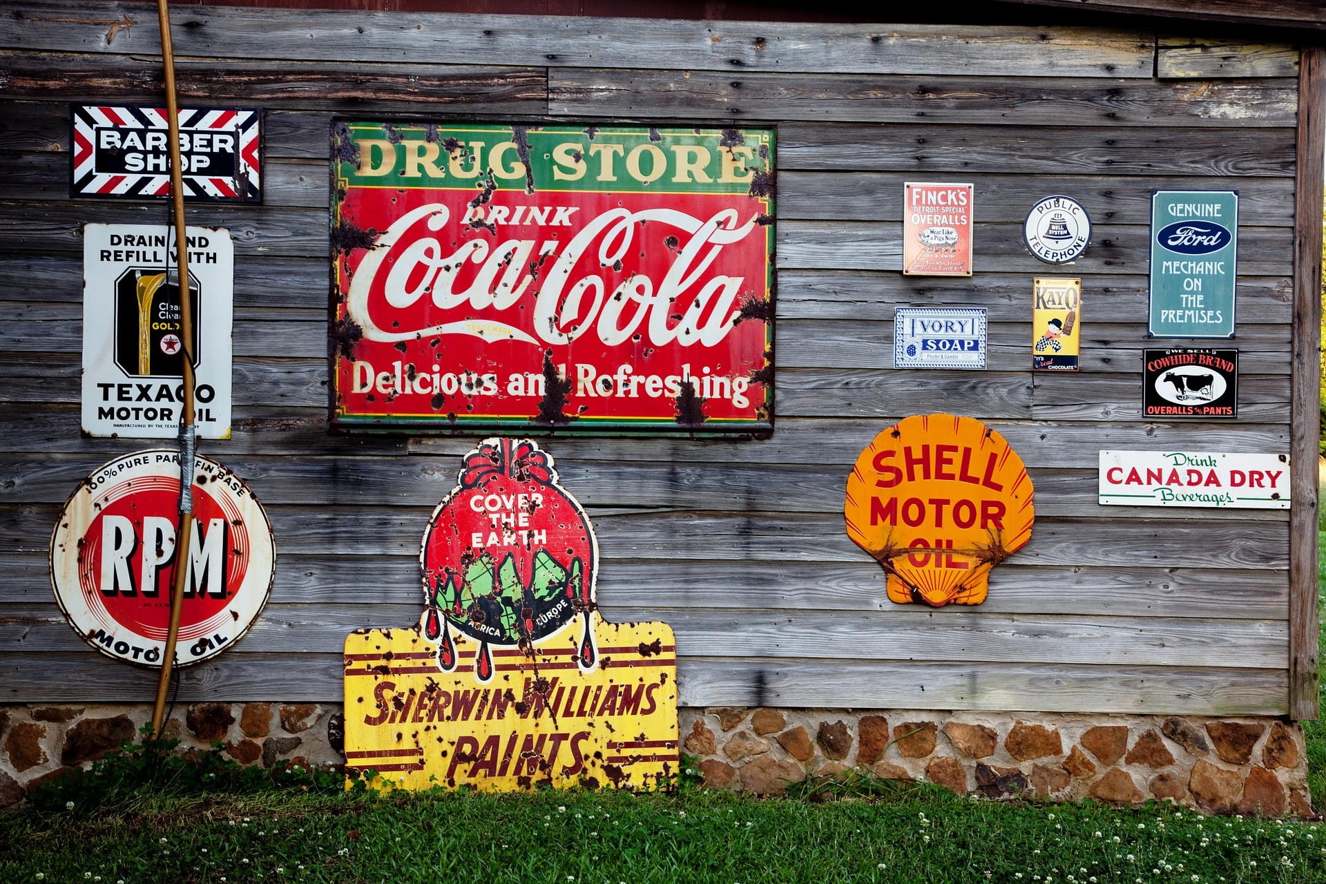 Auf einem Holzzaun sind sehr viele Werbeschilder von verschiedenen Marken wie Ford, Shell, Coca Cola und RPM Motor Oil befestigt.