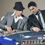 Zwei elegant gekleidete Männer sitzen an einem Pokertisch und besprechen sich.