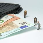 Neben einer offenen Geldbörse mit Euro-Scheinen stehen drei Männleins. Einer von ihnen zieht an einem Geldschein.