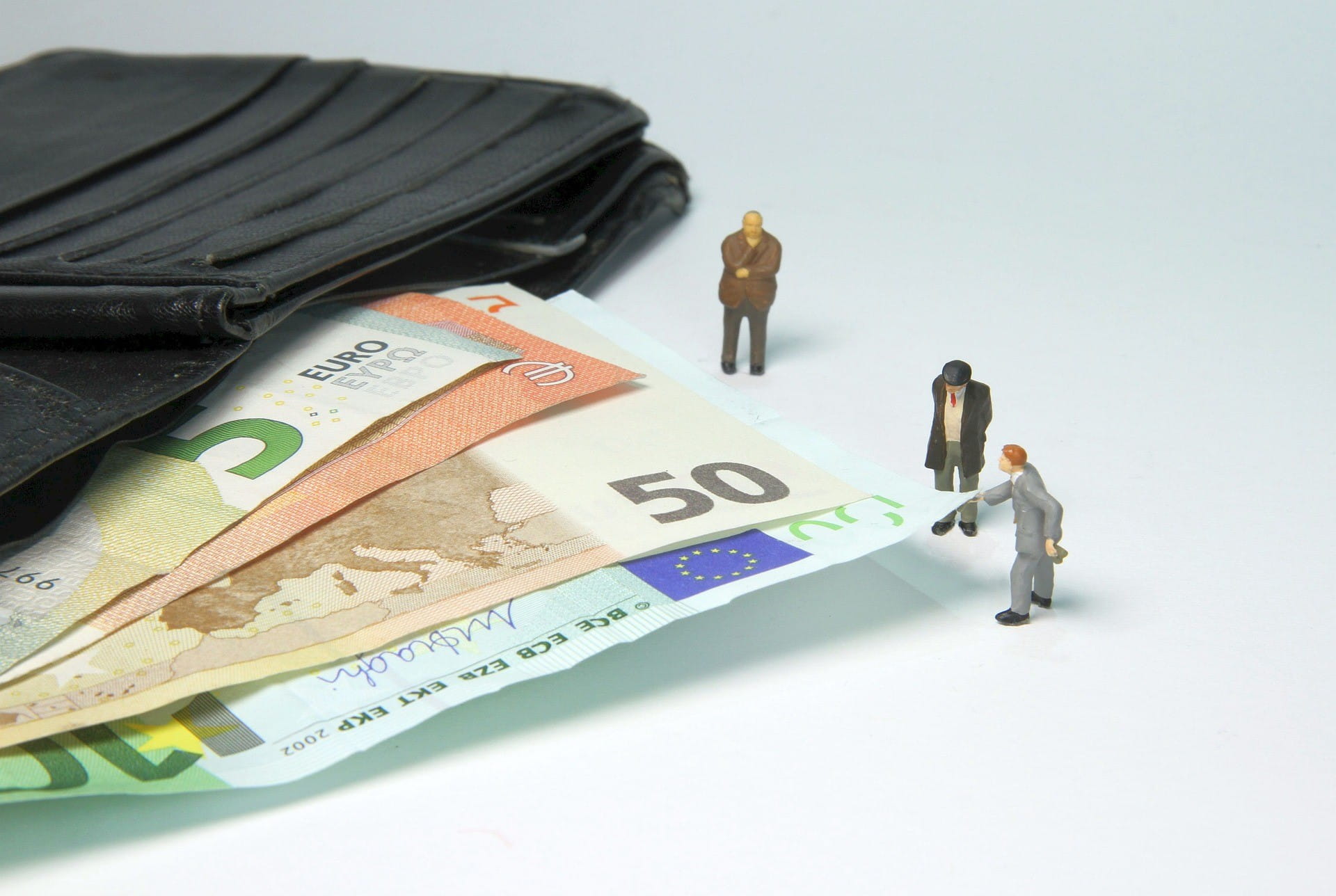 Tiga pria kecil berdiri di samping dompet terbuka dengan uang kertas euro.  Salah satunya menarik tagihan.
