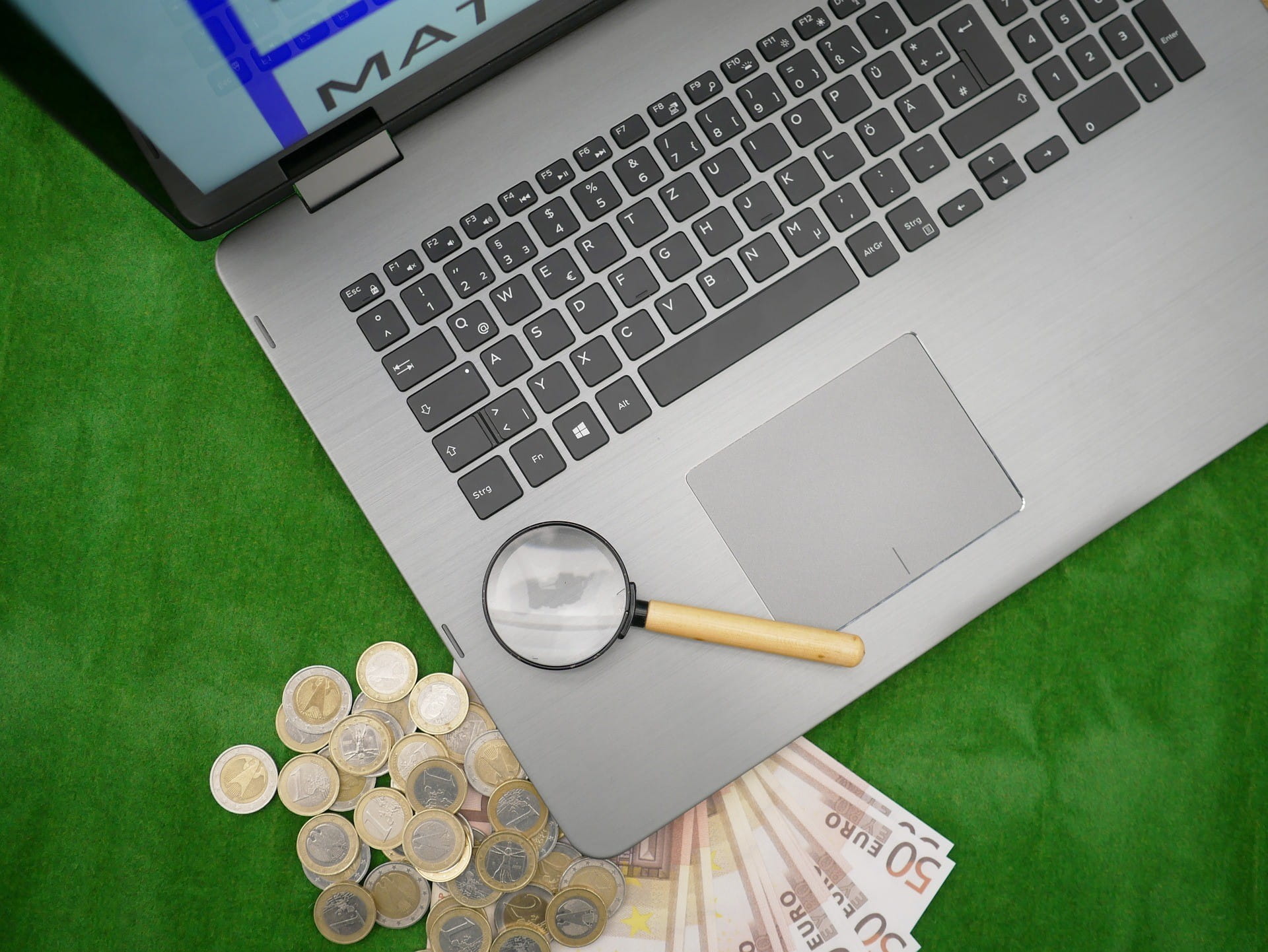 Ada uang kertas dan koin di bawah dan di samping laptop – ada kaca pembesar di laptop yang terbuka.