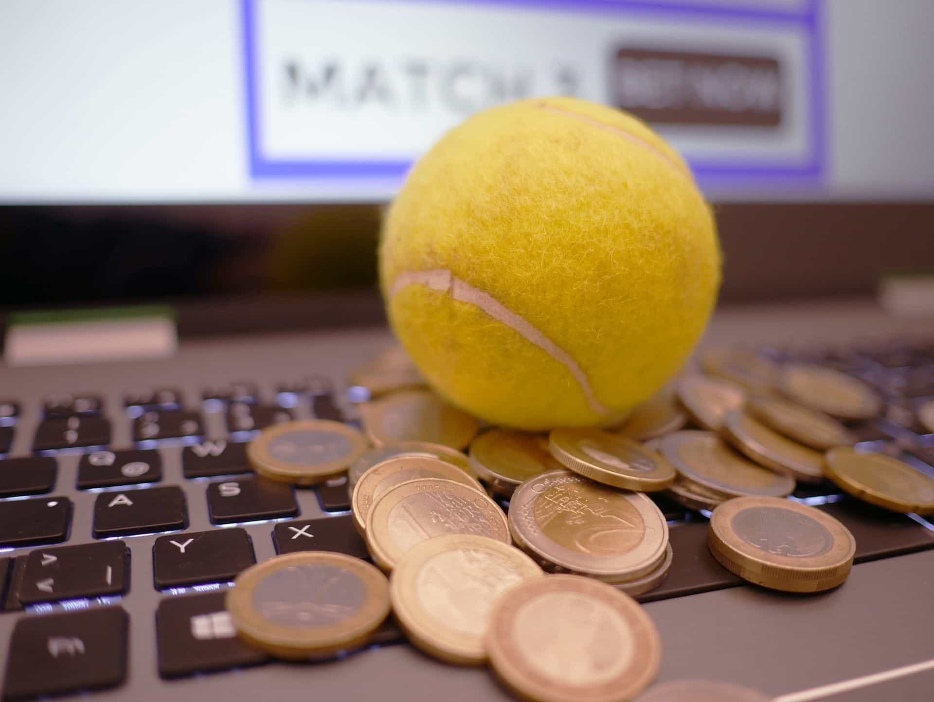 Auf der Tastatur eines Laptops liegen mehrere Euro-Münzen und ein gelber Tennisball.