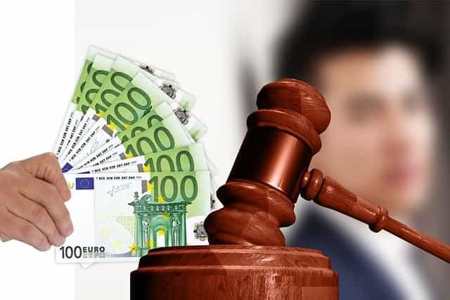 Links hält eine Hand mehrere 100-Euro-Scheine, während auf der rechten Seite ein Gerichtshammer auf der Holzplatte liegt - dahinter ist ein Mann.