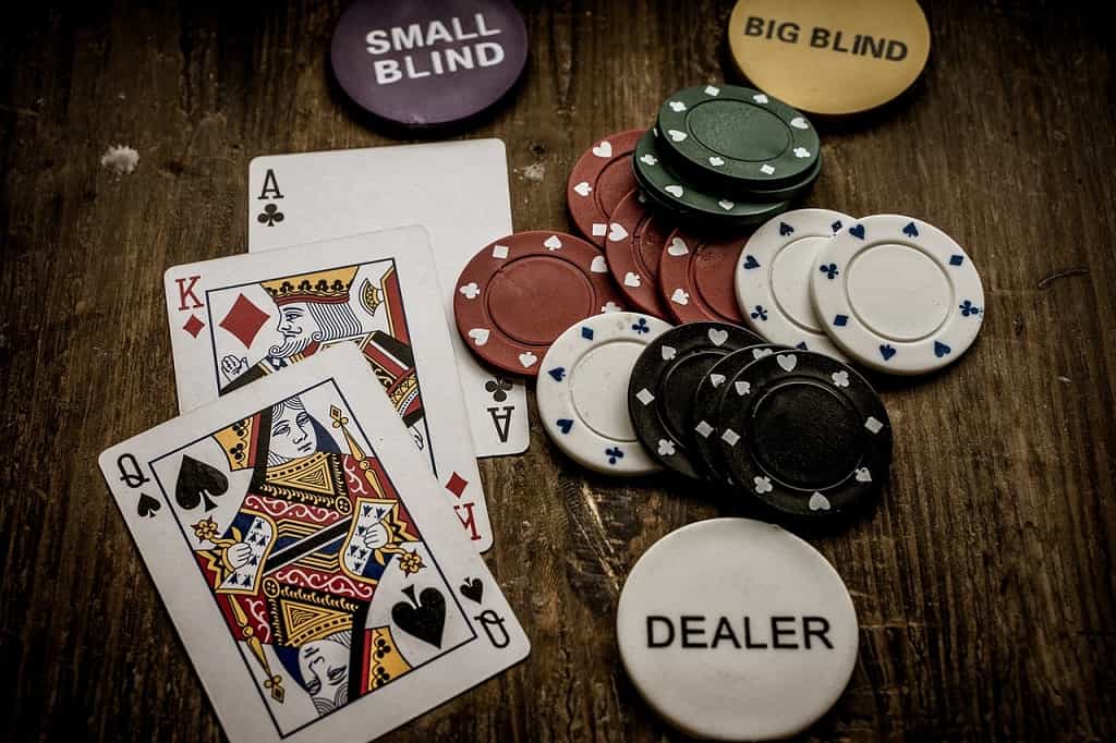 Di atas meja kayu ada tiga kartu remi, beberapa chip remi dan satu chip bertuliskan Dealer.
