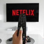 TV-Bildschirm, auf dem ein Netflix Logo zu sehen und uns eine Hand, die eine Fernbedieung hält.